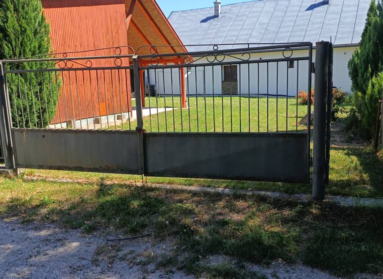 A gate and a wooden cottage in Tužina, surrounded by a lawn and coniferous trees.