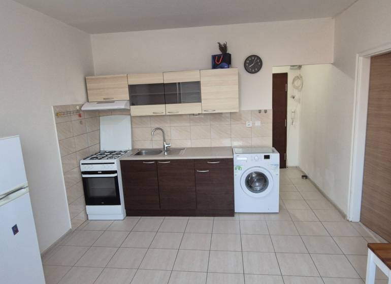 A kitchen in a studio apartment with a sink, stove, washing machine, and refrigerator on tiled flooring.