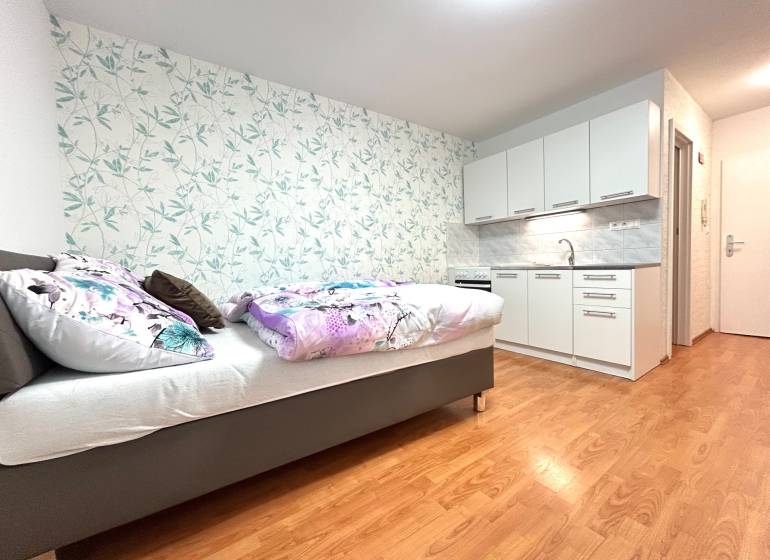 Studio apartment with a bed, kitchenette, and floral wallpaper on the wall.