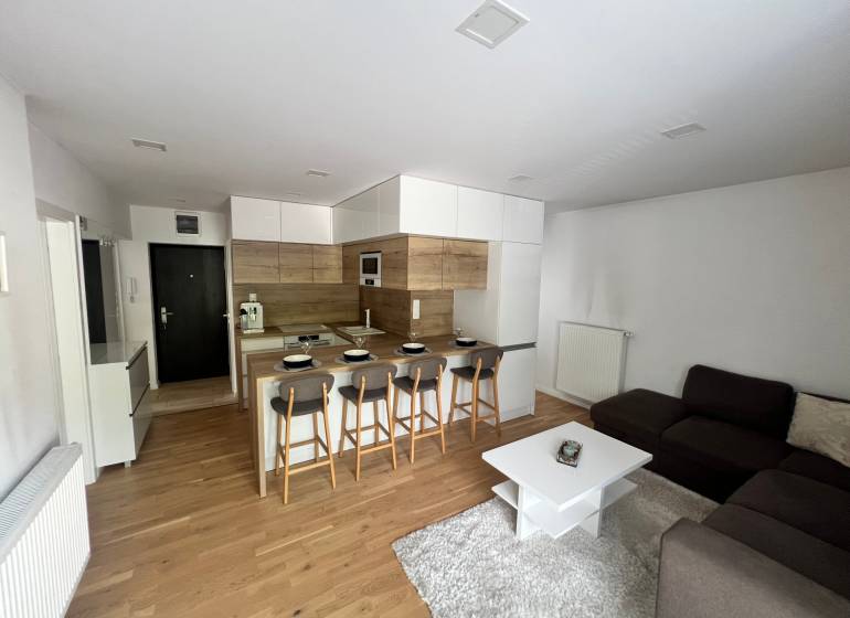 Living area of a 3-room apartment with an open kitchen, bar counter, and gray sofa.