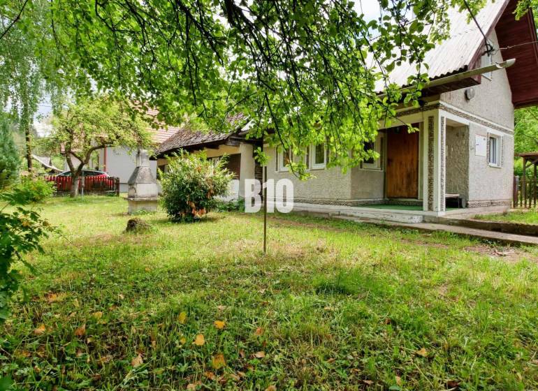 A cottage in Necpaly with a lawn and an outdoor fireplace, surrounded by greenery and trees.