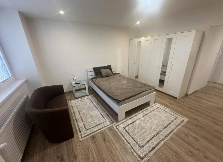 A room with a bed, a chair, and a wardrobe in a studio apartment, with a wooden decor floor.