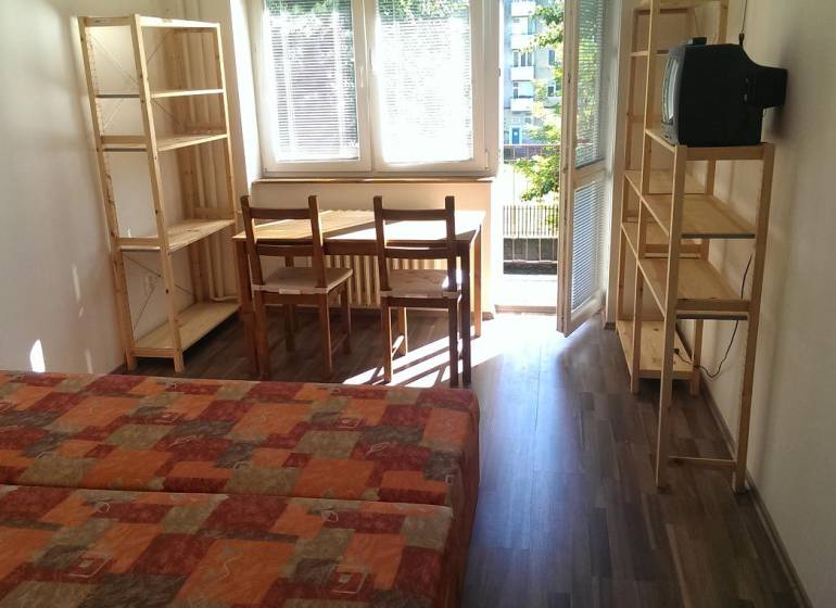 A one-room apartment with a wooden decor floor, a bed, a table, chairs, and shelves.