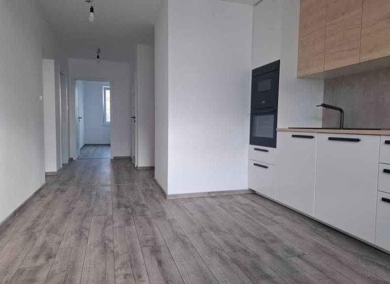 The kitchen of a 3-room apartment with white cabinets, a wooden decor floor, and a hallway.