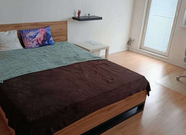 A bedroom in a one-room apartment with a wood-patterned floor, a bed, and an office chair.