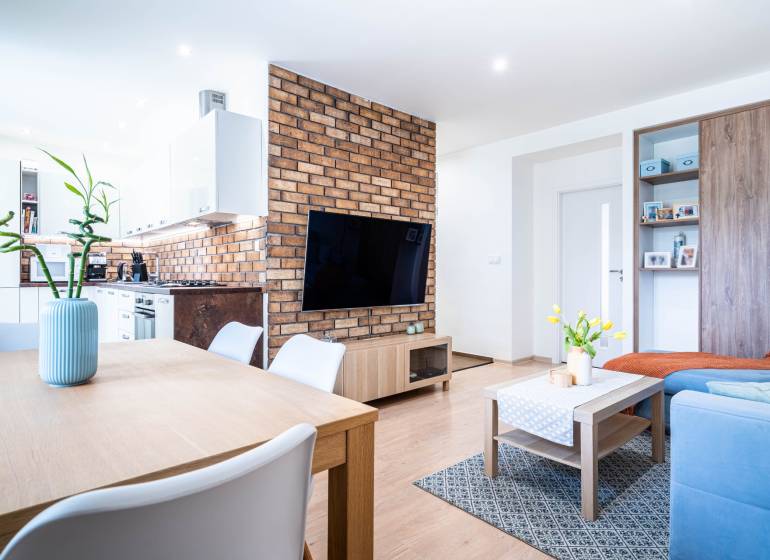 Living room with brick cladding, wood-patterned flooring, and a kitchen in a three-room apartment.
