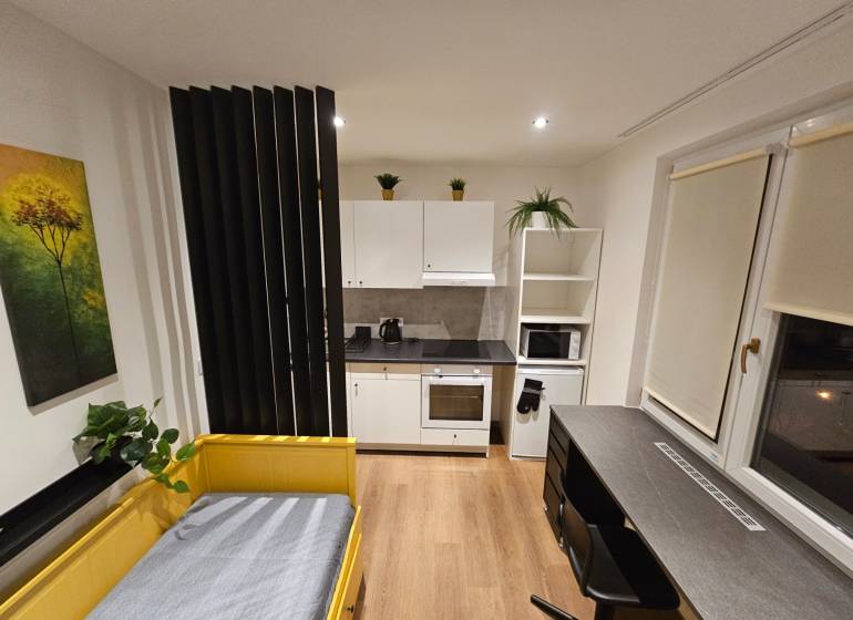 Studio apartment with a yellow bed, kitchen, table, and a wooden-patterned floor.