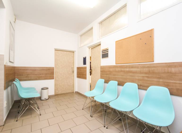 A waiting room with blue chairs, a cork bulletin board, and a wooden-patterned floor in an office.