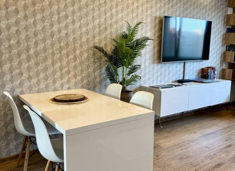 Dining table with white chairs in a 4-room apartment, wooden floor, TV on the wall.