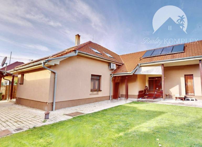 A family house in Komárno surrounded by a green lawn and paving under a blue sky.