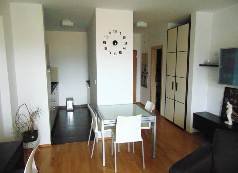 Interior of a 1-bedroom apartment with a dining table, wood-patterned flooring, and a clock on the wall.