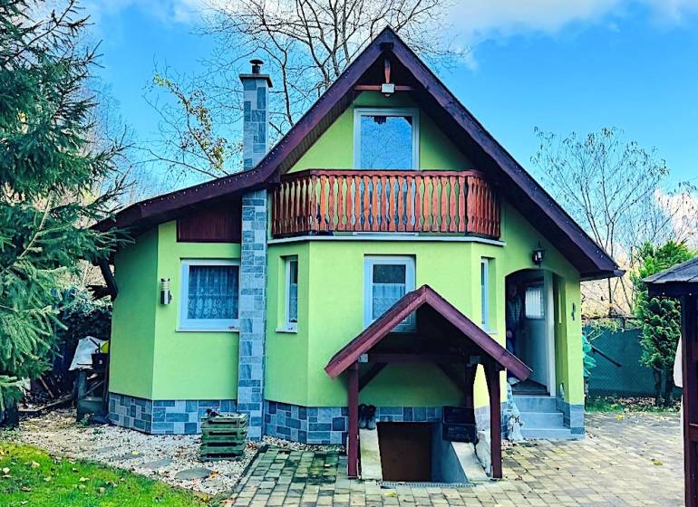 A cottage in Partizánske with a wooden balcony, surrounded by greenery and a paved courtyard.