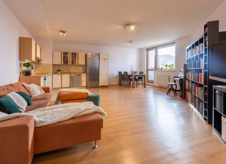 Living room in a three-room apartment with a wood-decor floor, corner kitchen, and dining table.