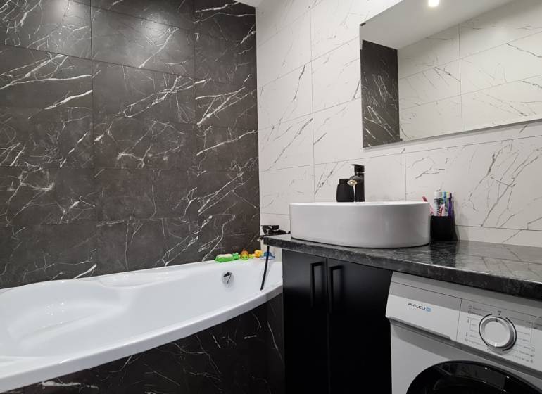 A bathroom with a bathtub, sink, and washing machine in a 2-room apartment, tiled with dark marble tiles.