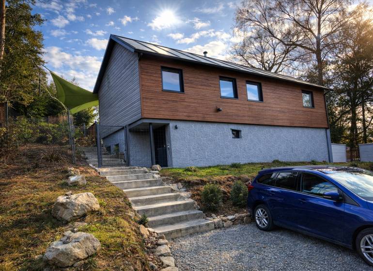A cottage in Zvolen with wooden cladding, stairs, and a parked car on a gravel surface.