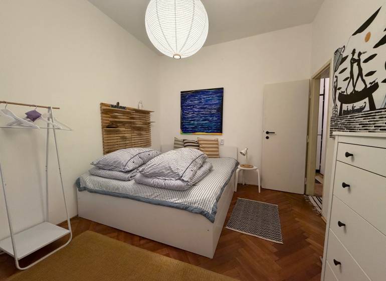 Bedroom in a two-room apartment with wood-patterned flooring, a bed, and a white cabinet.