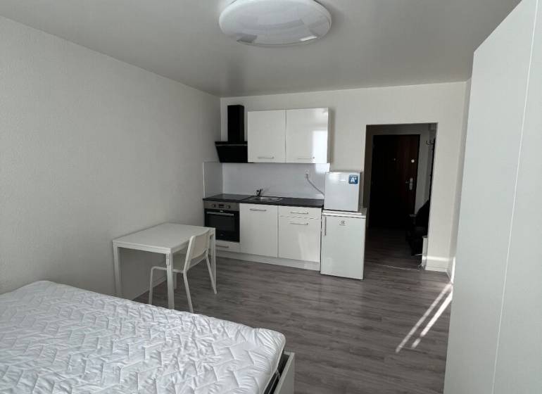 Studio apartment with a white kitchen unit and wood-patterned flooring, bed, and table.