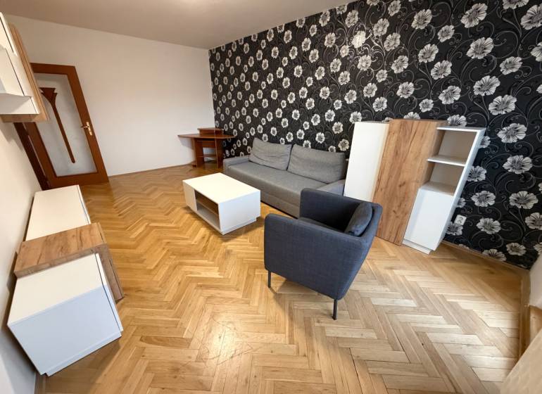 Living room in a three-room apartment with a wooden-patterned floor, patterned wallpaper, and furniture.