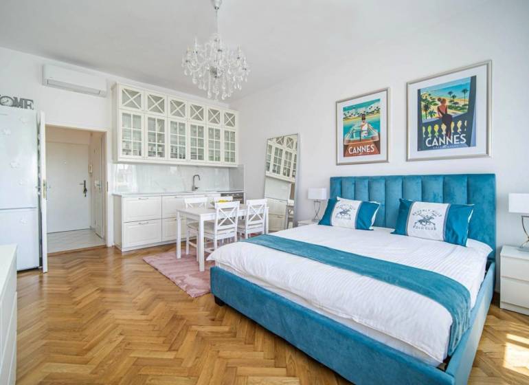 A one-bedroom apartment with a wood-patterned floor, a bed, and white kitchen cabinets.