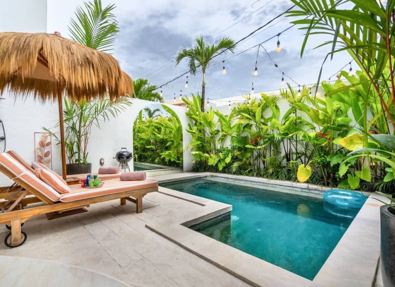 A villa pool with loungers, a rattan roof, surrounded by tropical plants.