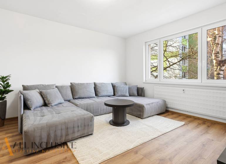 A bright living room with wood-patterned flooring, a gray sofa, and a window in a three-room apartment.