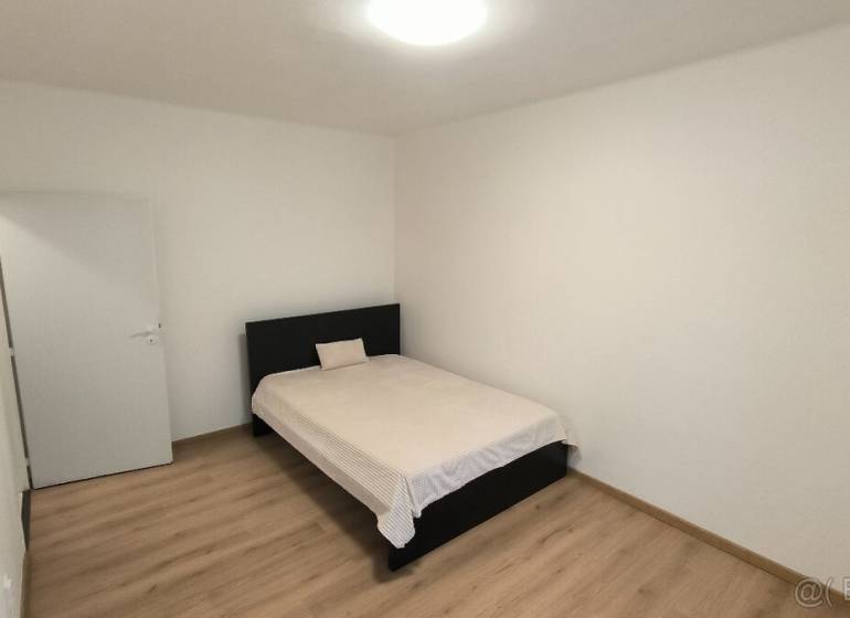 A bedroom in a studio apartment with a bed, white walls, and a floor with a wooden decor.