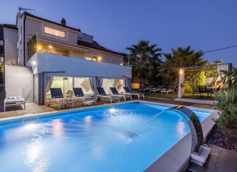 Villa in Kaštela with a lit pool, loungers, and palm trees in the garden at dusk.