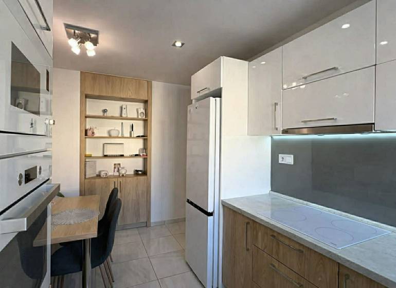 A kitchen in a 2-room apartment with white cabinets, a table, and wooden decor shelves.