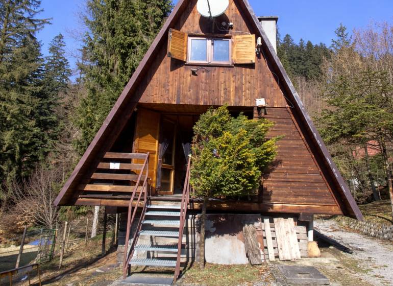 A wooden cottage in Oščadnica surrounded by greenery and conifers in mild sunlight.