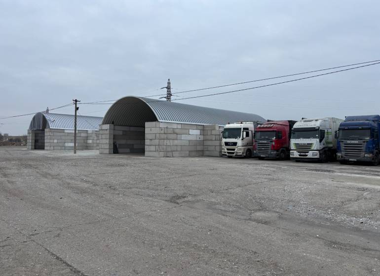 Warehouses and halls in Ladce with parked trucks on a gravel parking lot.