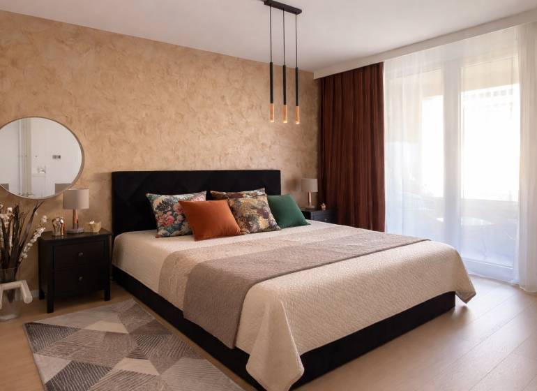Bedroom in a two-room apartment with a large bed, mirror, and wooden decor flooring.