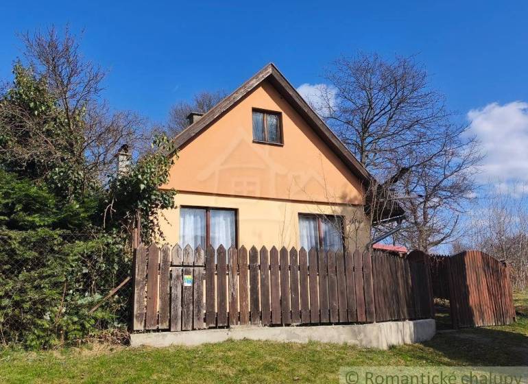 A cottage in Hronská Dúbrava surrounded by greenery and a wooden fence, with a clear blue sky.