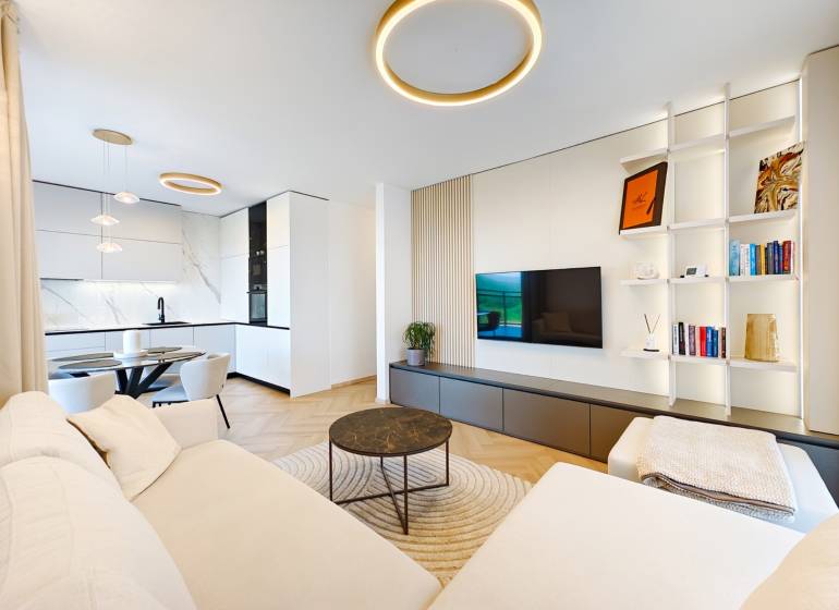 Living room of a two-room apartment with a sofa, kitchen, and wooden decor flooring.