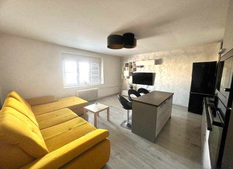 Living room with a large yellow sofa, kitchen island, and television in a 2-room apartment.