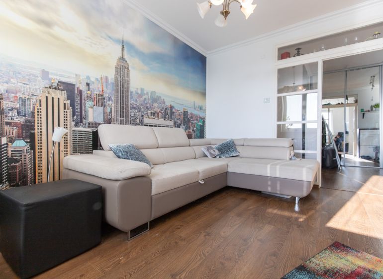 Living room in a 2-room apartment with a beige sofa, city photo wallpaper, wooden decor flooring.