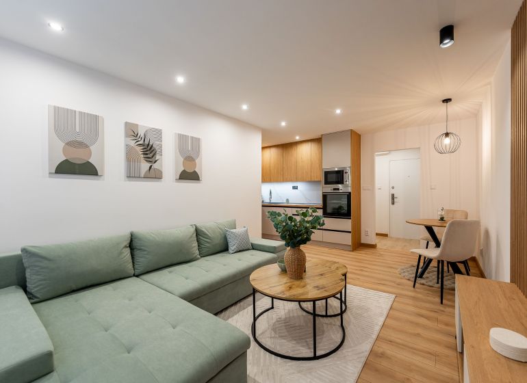Living room of a two-room apartment with furniture, kitchen, and wooden decor flooring.
