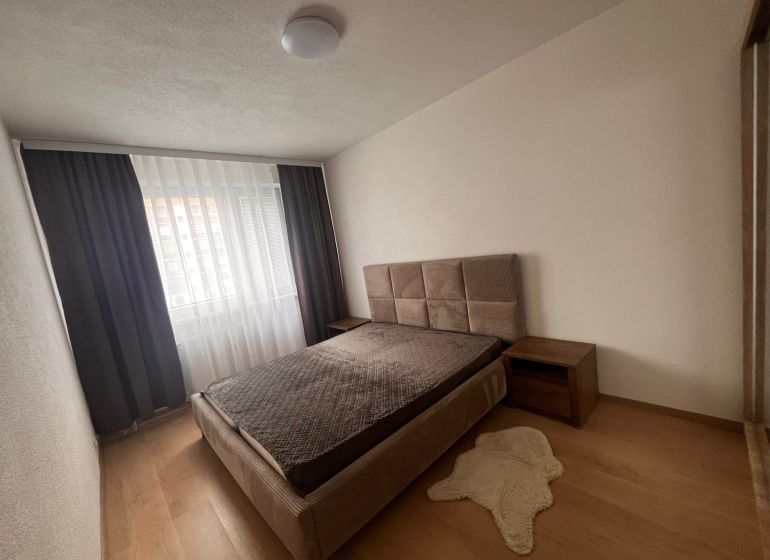 A bedroom in a 2-room apartment with a wooden decor floor and a bed by the window.