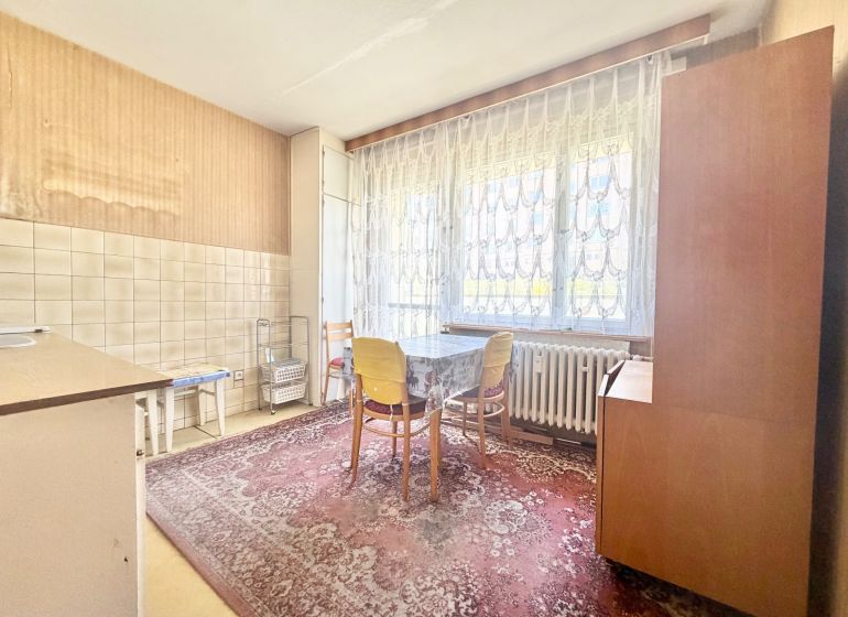 A kitchen in a 2-room apartment with a table, a carpet, and a large window with lace curtains.