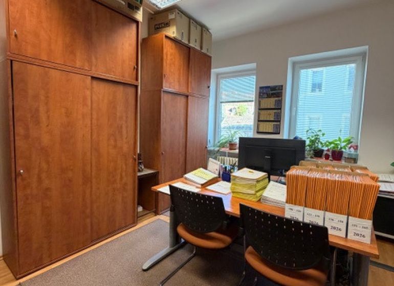 Offices with wooden furniture, documents on the table, two windows, and hanging ceiling lights.