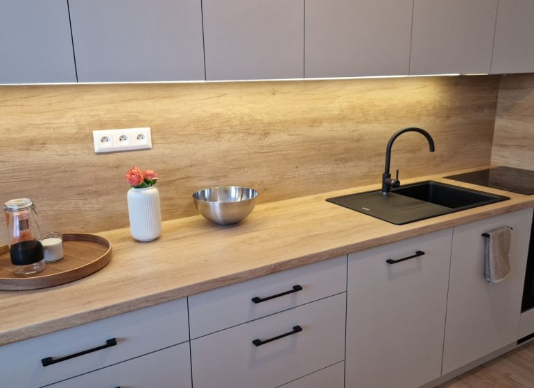 A kitchen in a 3-room apartment with a wooden decor floor, light cabinets, and a black sink.