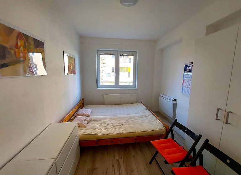 Studio apartment with a bed, white wardrobe, wood-patterned flooring, two folding chairs.