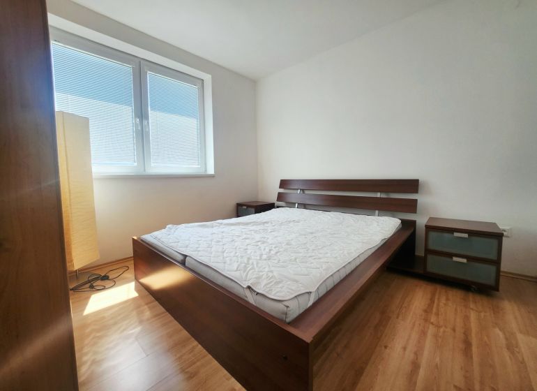 A bedroom with sunlight, a bed, a nightstand, and a wooden-patterned floor.