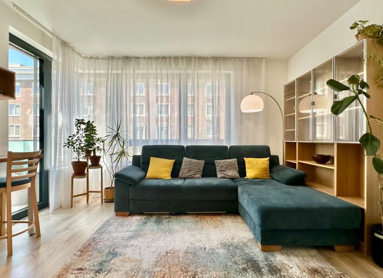 Living room in a 2-room apartment with a couch, shelves, plants, and a floor with a wooden decor.