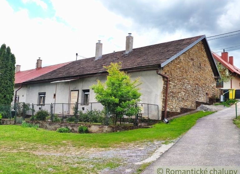 A cottage in Hermanovce with a stone wall and a garden by an asphalt road.