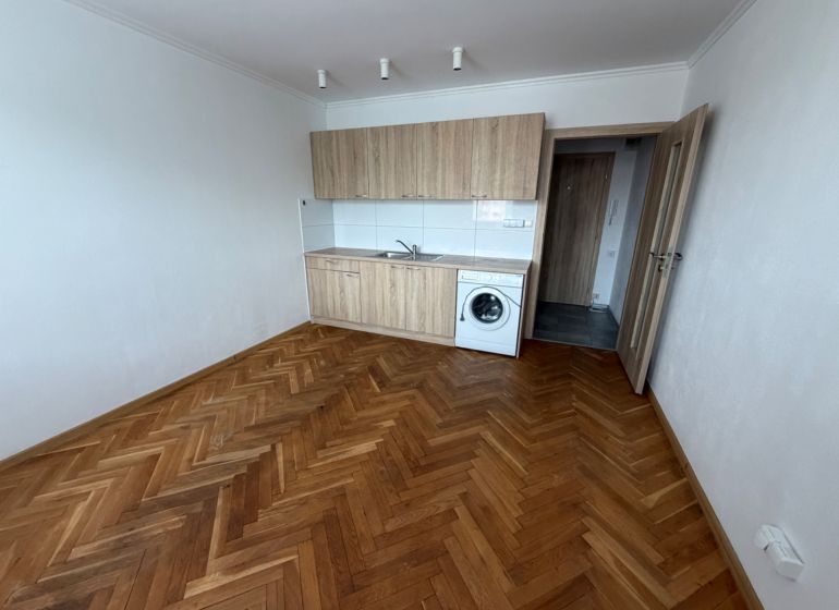 Studio apartment with a kitchenette, washing machine, and wooden decor flooring.