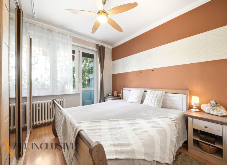 A bedroom in a 3-room apartment with a wood-patterned floor, a fan, and balcony windows.