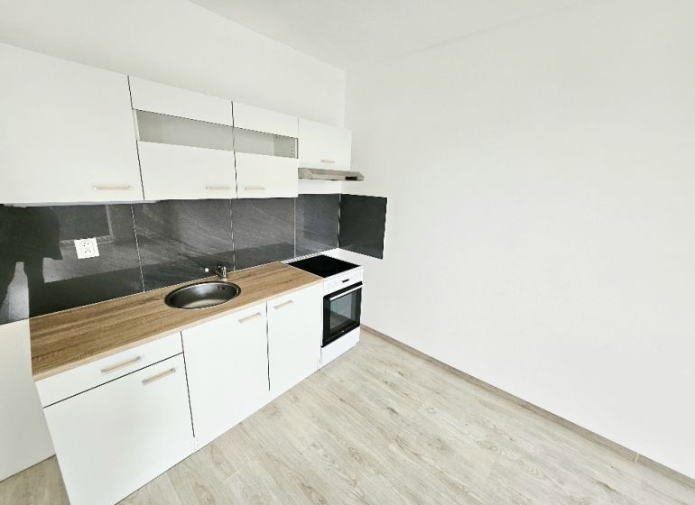 White kitchen unit with black tiles in a studio apartment with a wood-patterned floor.