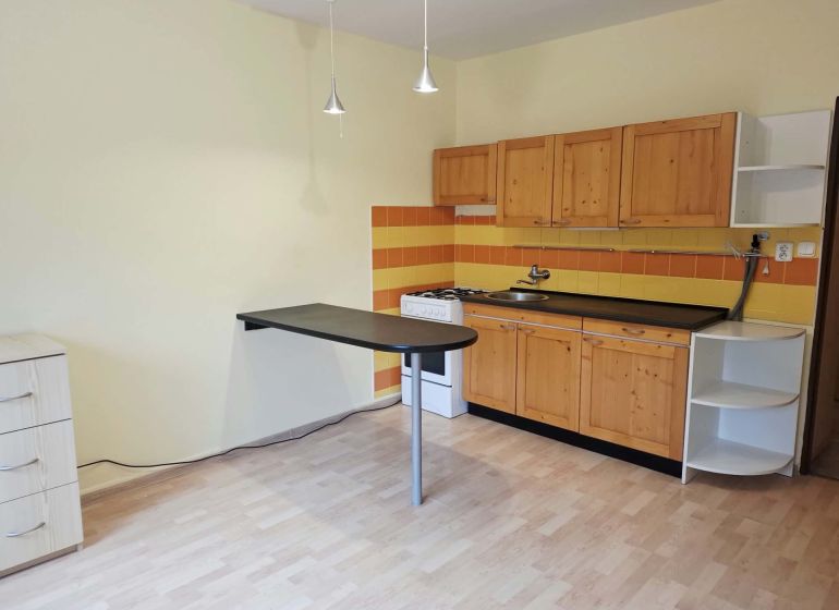 A kitchen in a one-room apartment with a wooden decor floor, wooden cabinets, and orange tiles.
