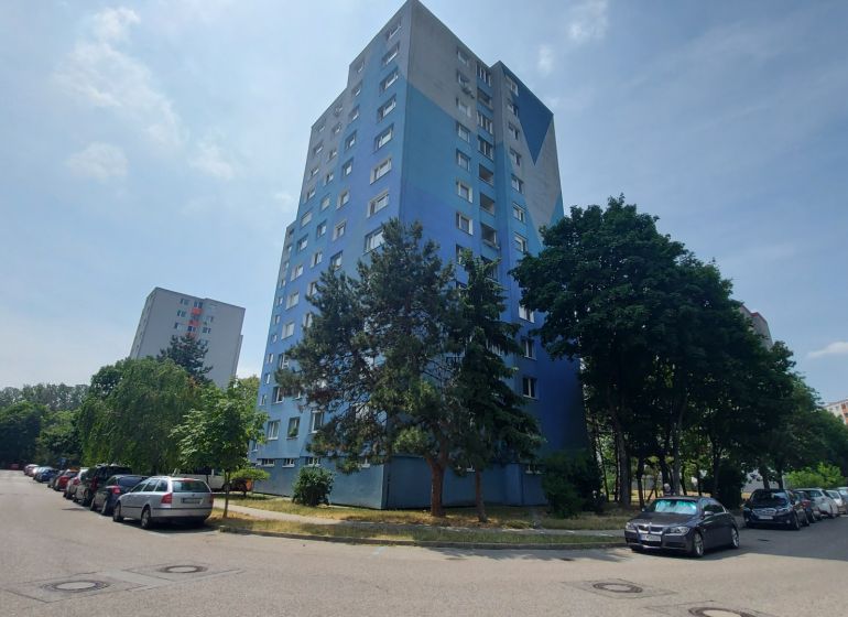 A taller residential apartment building in Bratislava - Petržalka surrounded by greenery and parked cars.
