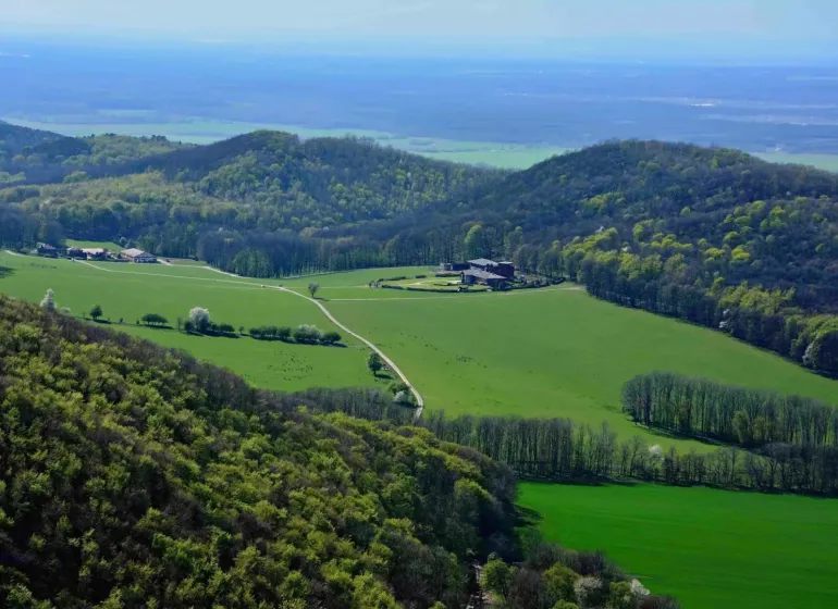 Green agricultural and forest lands around Plavecký Peter with hills and a farm.